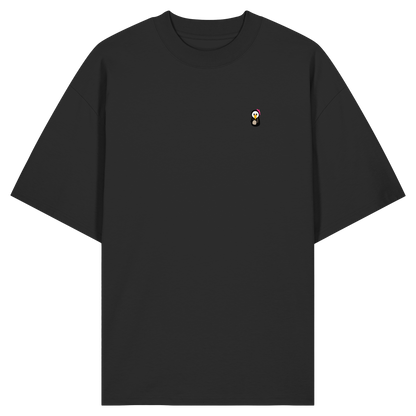 Die Vorderansicht eines schwarzen Oversized-T-Shirts mit klassischem Rundhalsausschnitt und weiten Ärmeln. Auf der linken Brust befindet sich ein kleiner, minimalistischer und kontrastreicher Print eines Pinguin-Charakters mit großen Augen, gelbem Schnabel und einer leuchtend pinken Schleife am Kopf. Das Produktbild ist vor einem transparenten, karierten Hintergrund freigestellt